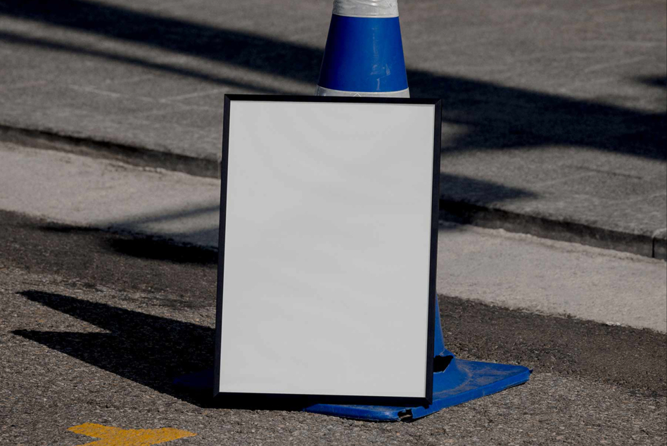 Blank white poster frame behind blue cone on asphalt with strong sunlight shadows