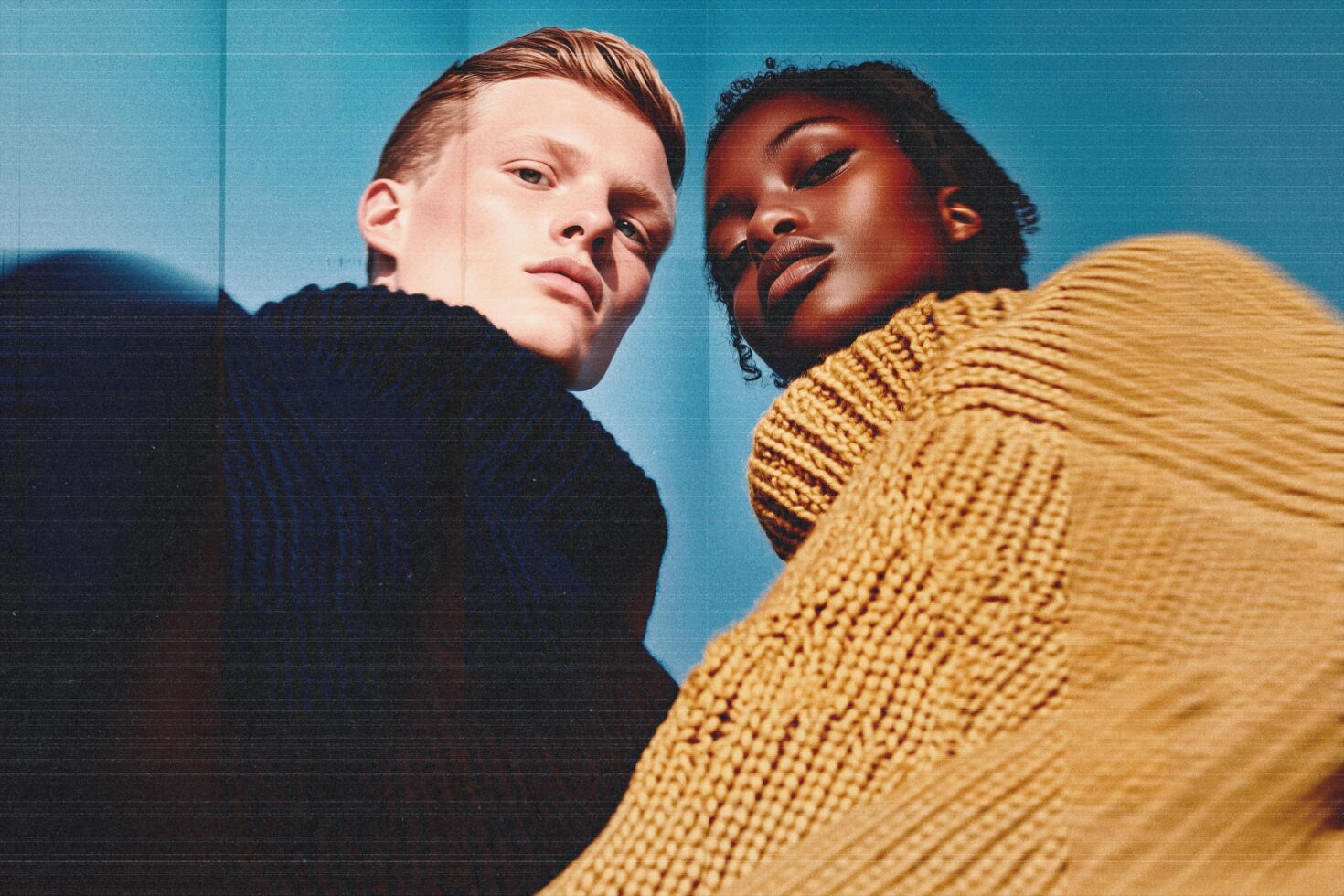 Two people against blue background wearing black textured turtlenecks looking away