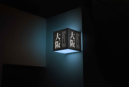 Cube-shaped corner sign with white Japanese characters on dark panels, blue interior glow