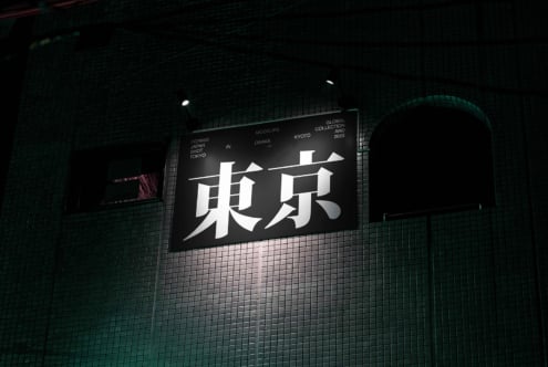 Illuminated horizontal billboard with large white Japanese characters on dark background, teal brick wall