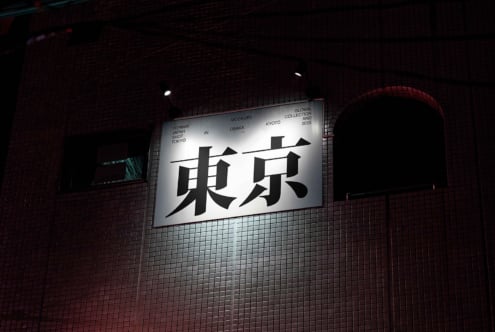 Illuminated horizontal billboard with bold black Japanese characters on light background, brick wall with red tones