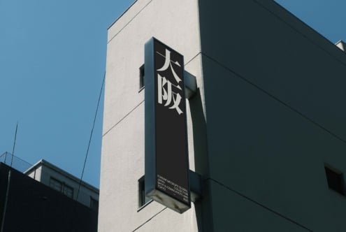 Vertical black metal sign with white kanji on light building facade, blue sky