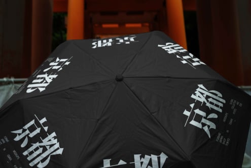 Black umbrella with white Asian characters printed across panels, opened in front of orange architecture