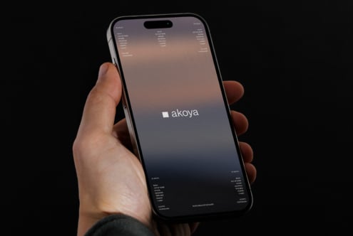 Hand holding iPhone against dark background displaying Akoya site with soft gradient design