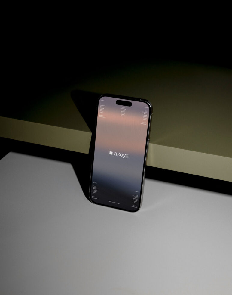 iPhone propped on geometric surface showing Akoya website with peach gradient design
