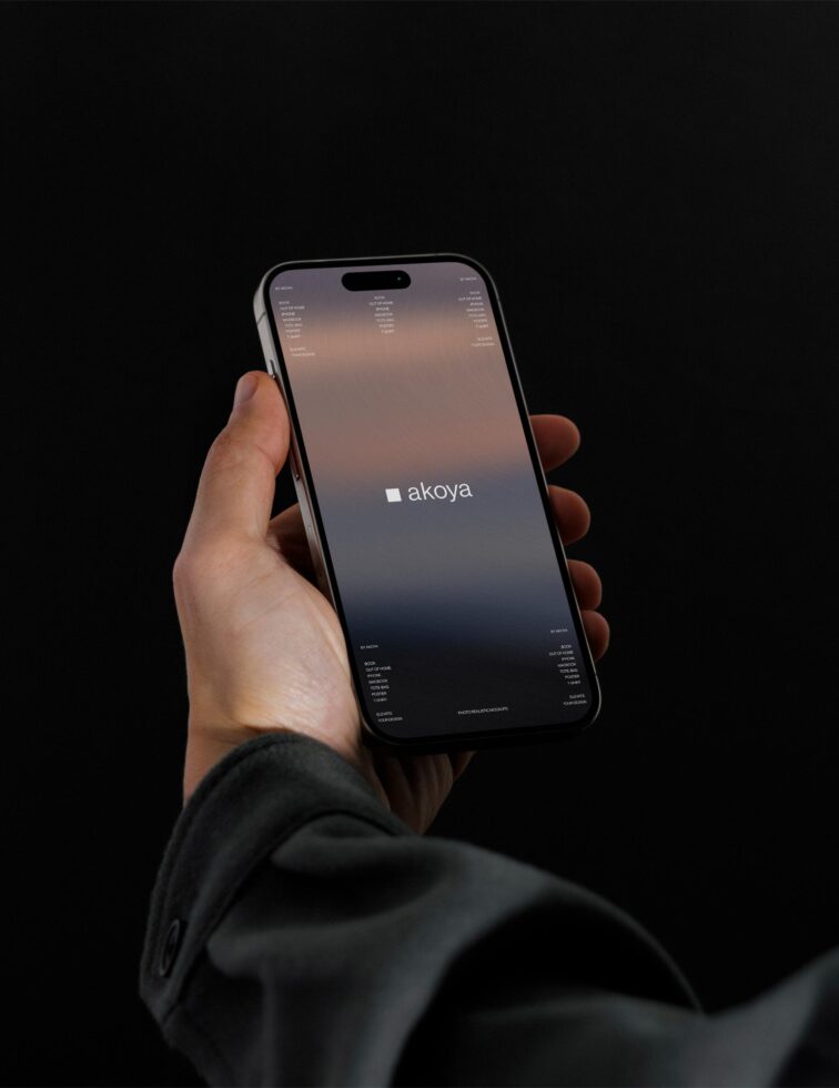 Hand holding iPhone showing Akoya website with sunset gradient screen against dark backdrop