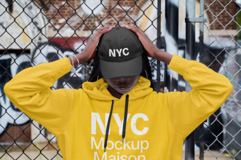 Person in bright yellow hoodie adjusting black NYC cap against chain-link fence