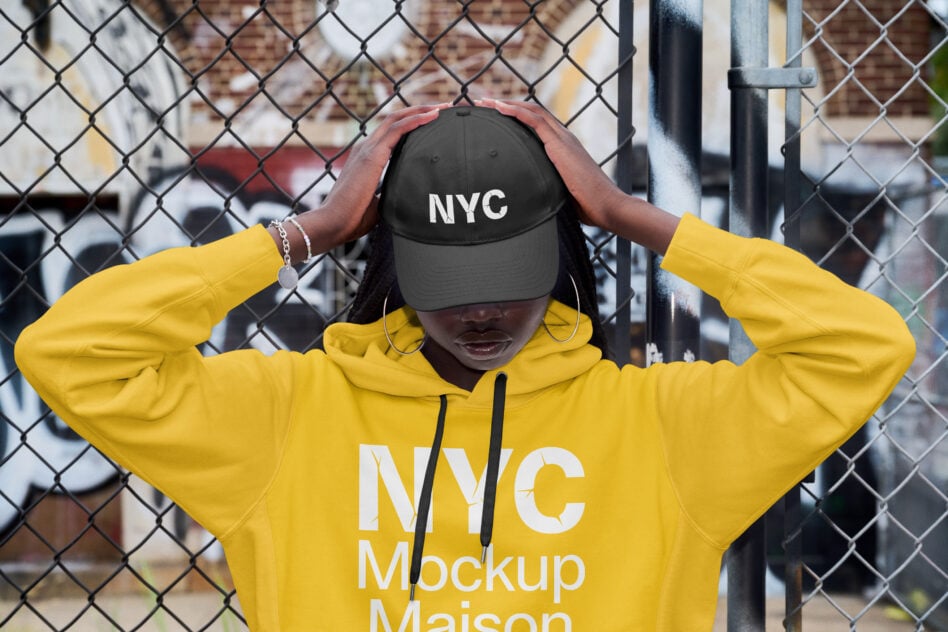 Person in bright yellow hoodie adjusting black NYC cap against chain-link fence