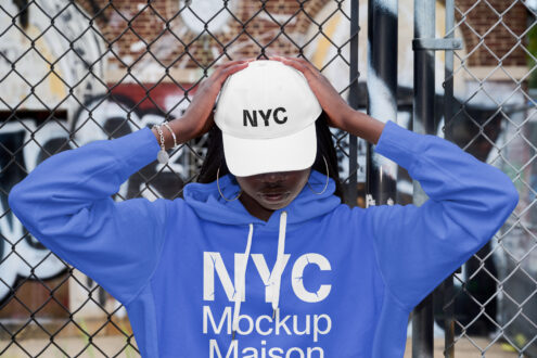 Woman by chain fence holding white cap wearing blue hoodie streetwear