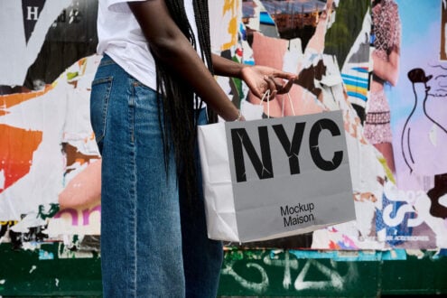 Hand holding white tote bag with black NYC logo against street mural