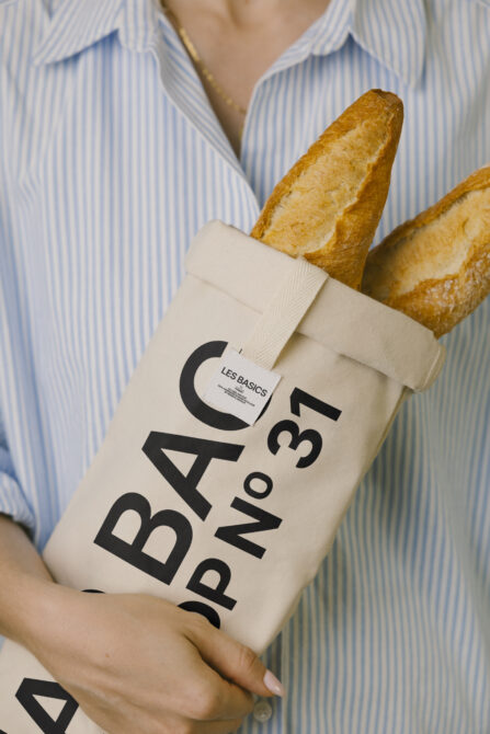 Cream colored fabric bag with Les Basics lettering held by someone, golden baguettes sticking out top