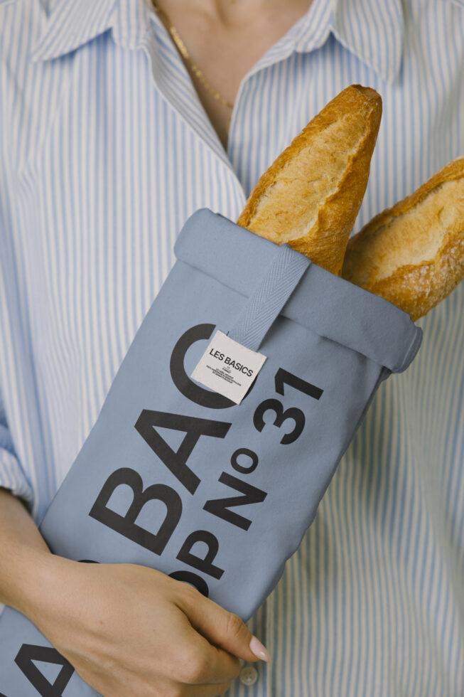 Person in striped shirt holding blue fabric bag with bold Les Basics branding, fresh baguettes tucked inside