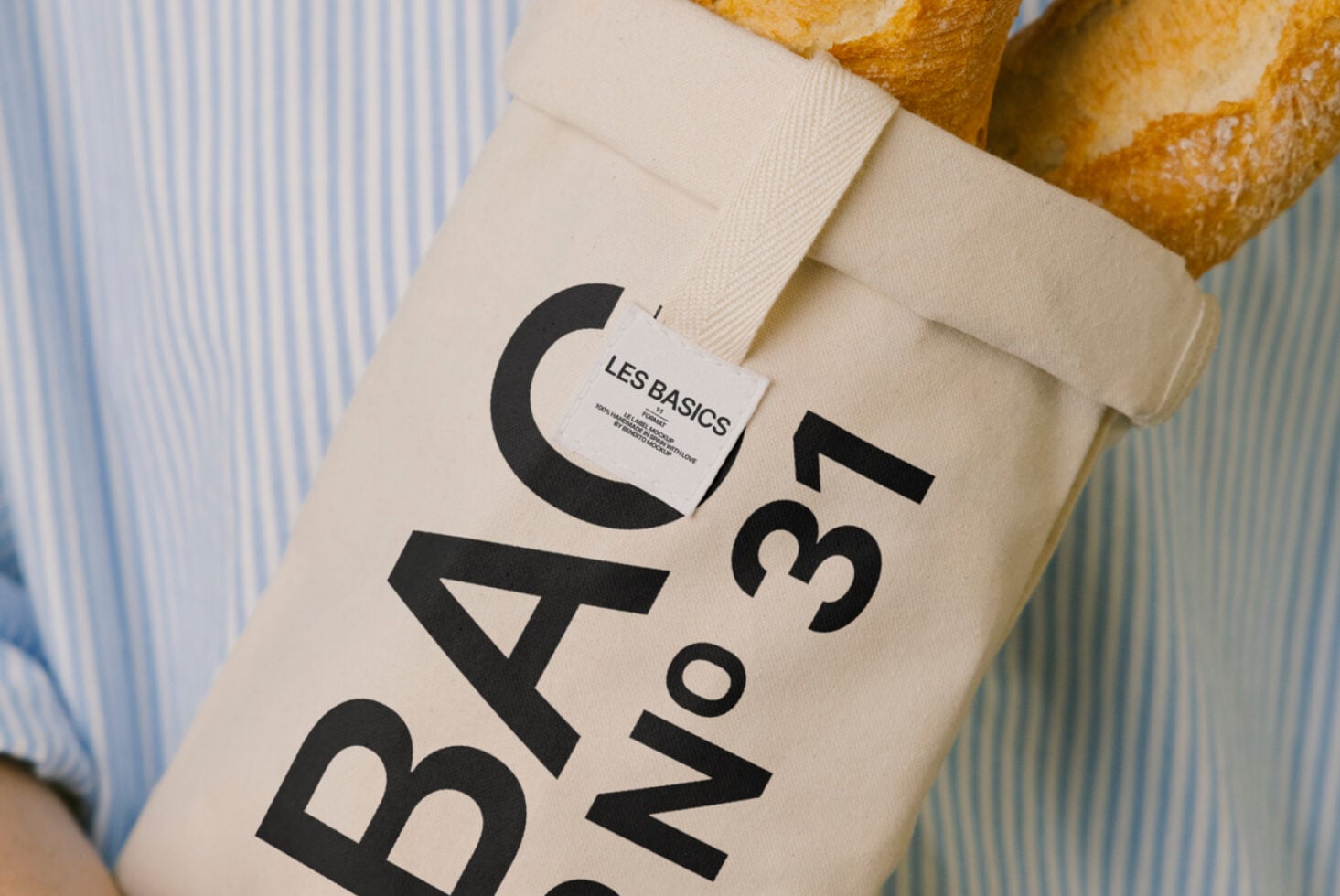 Cream fabric bag with bold Les Basics text mockup held against striped shirt, bread loaves visible