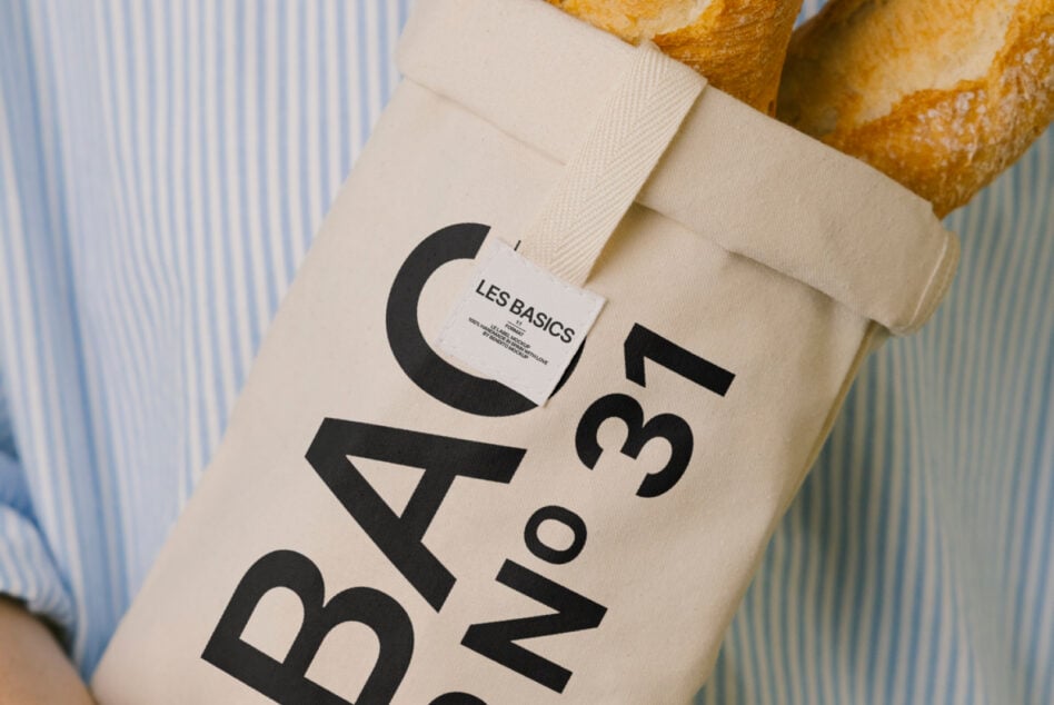 Cream fabric bag with bold Les Basics text mockup held against striped shirt, bread loaves visible