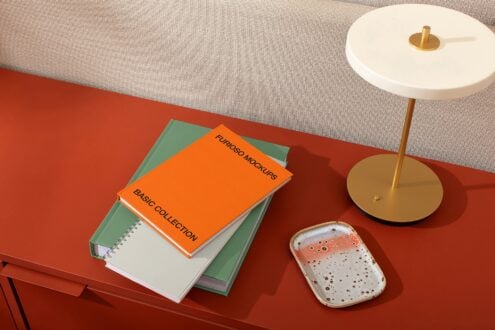 Stacked books with orange cover on red desk next to brass lamp, ceramic tray, neutral sofa backdrop