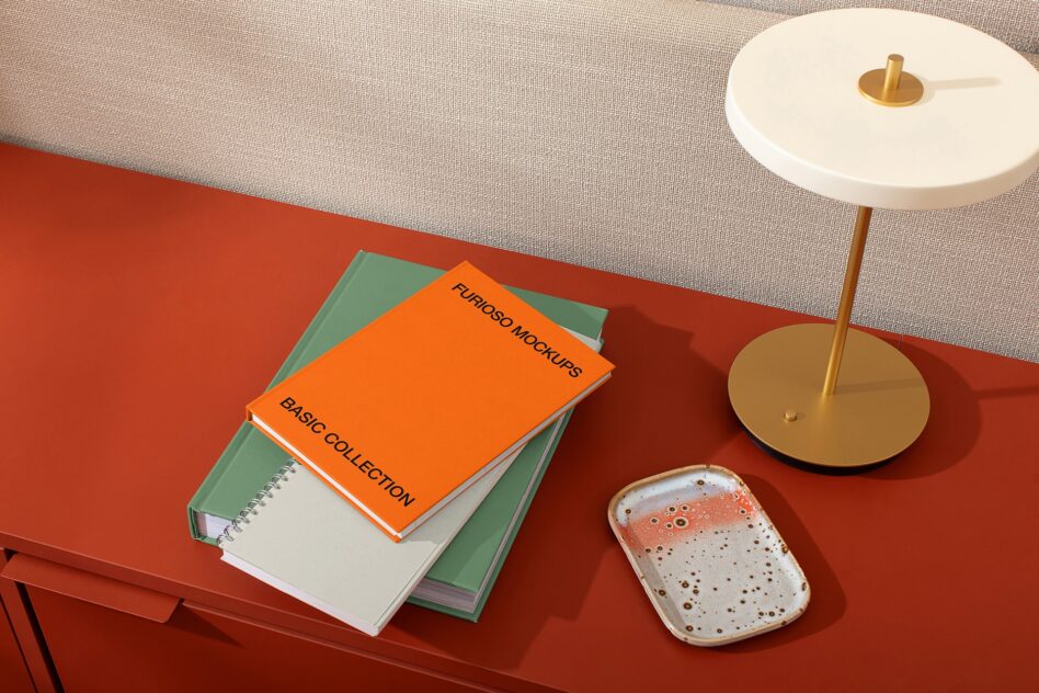Stacked books with orange cover on red desk next to brass lamp, ceramic tray, neutral sofa backdrop