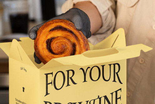 Gloved hand holding fresh pastry inside yellow branded box with text