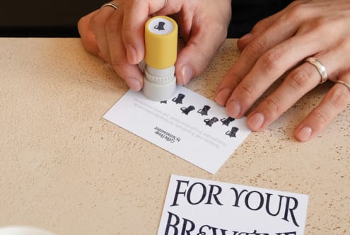 Yellow stamp with cat logo being pressed onto coffee order card on cork surface