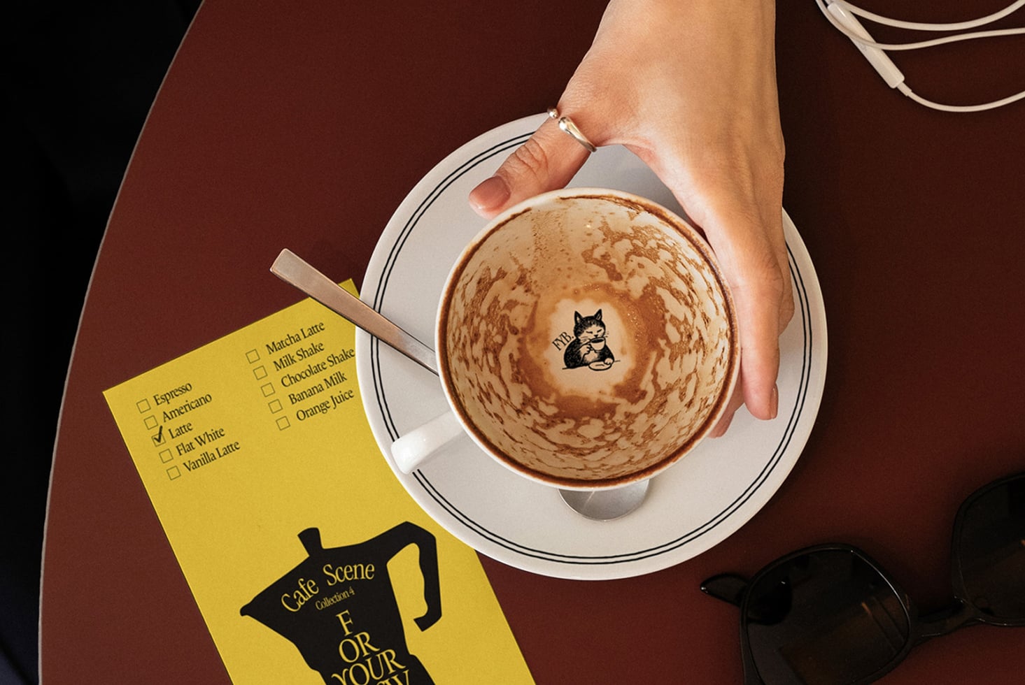 Overhead of latte with cat latte art, yellow coffee order card, sunglasses on burgundy