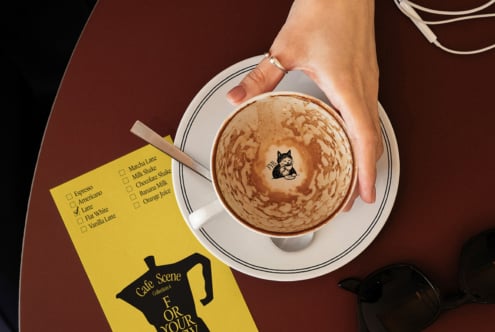 Overhead of latte with cat latte art, yellow coffee order card, sunglasses on burgundy