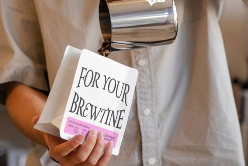 Woman in light shirt holding white coffee bag with bright pink label detail, pitcher tilted