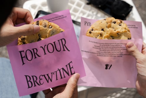 Two hands holding pink cookie bag mockups with chocolate chip baked goods inside