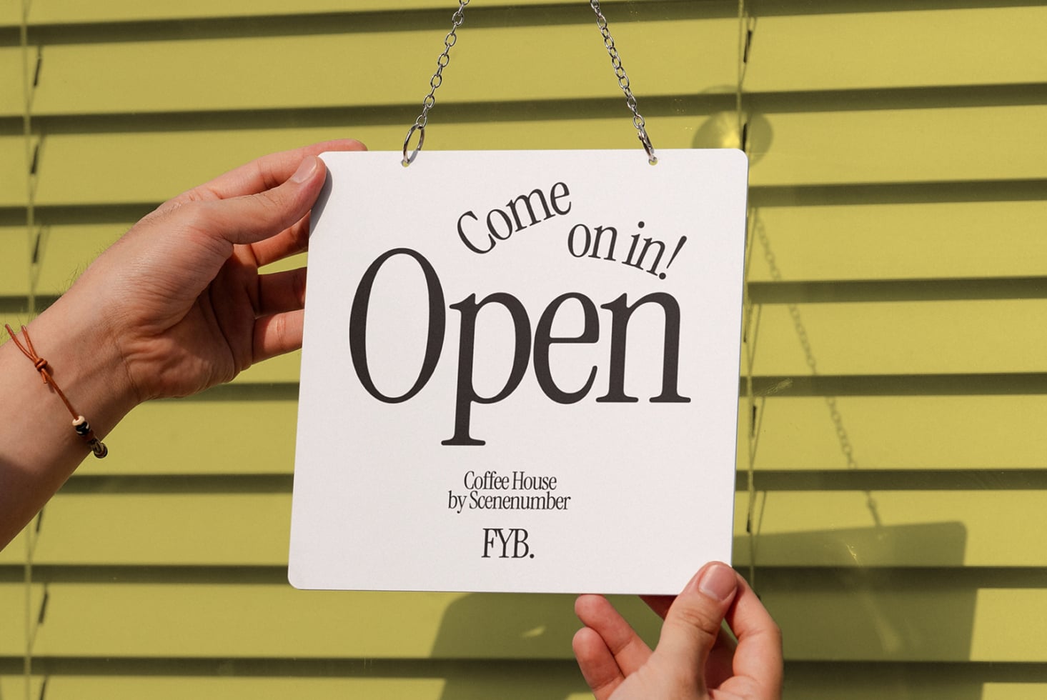 Hand holding Come on in Open door sign against yellow horizontal blind slatted wall