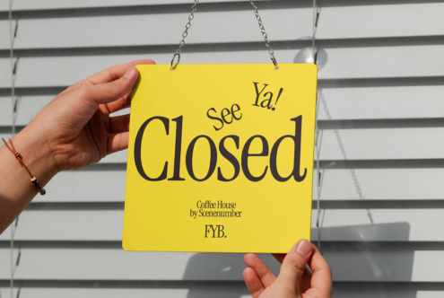 Hand holding bright yellow Closed see ya door sign against white horizontal blind backdrop