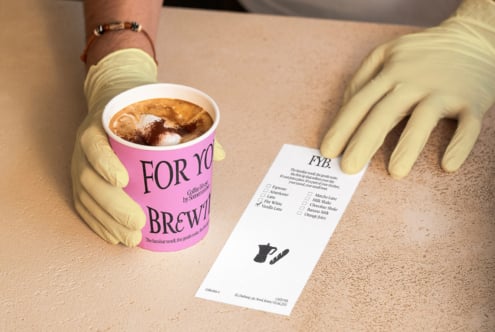 Pink coffee cup with latte art held in yellow gloved hands next to white menu card
