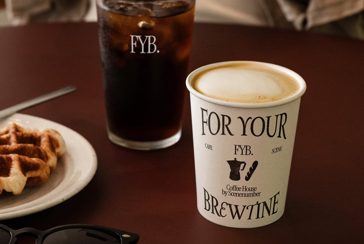Cream-colored coffee cup with latte next to iced cola in branded glass, pastry on plate