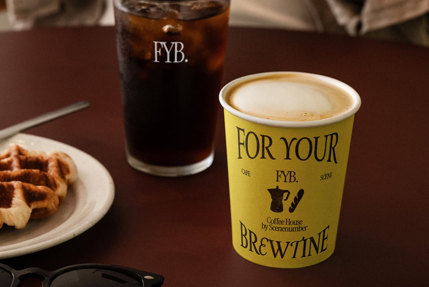 Yellow paper cup with latte next to clear glass with iced drink, pastry