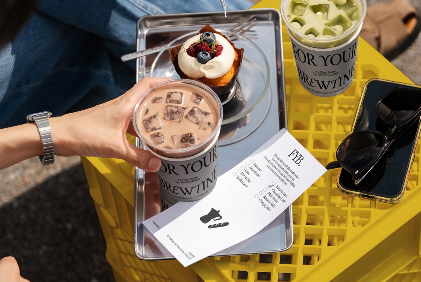 Branded paper cups held outdoors with chocolate pastry, yellow plastic crate, notepad