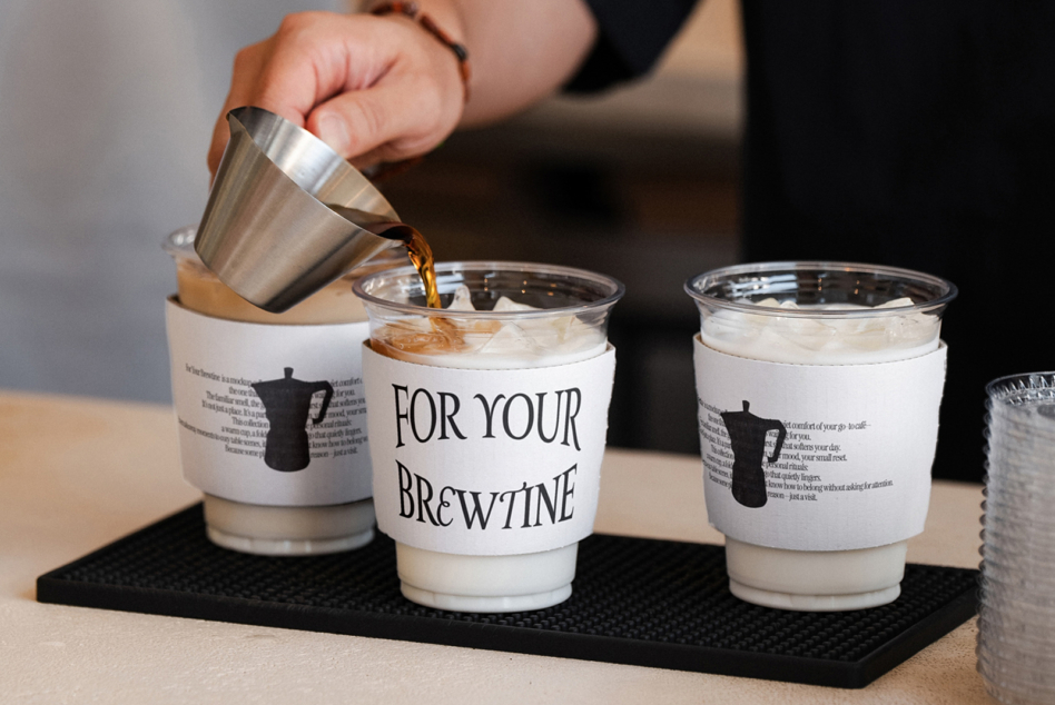 Barista pouring espresso into milk pitcher over three branded cups on black mat