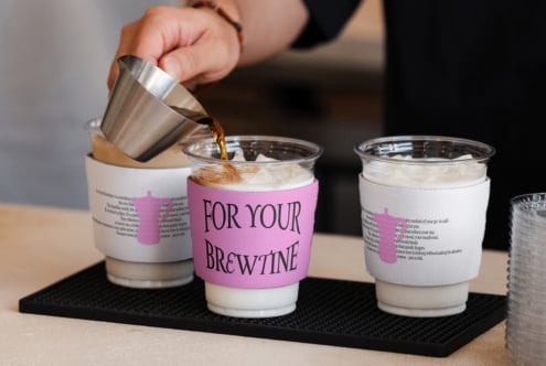Barista pouring espresso into cup with pink For Your Brewtine sleeve, three cups on tray