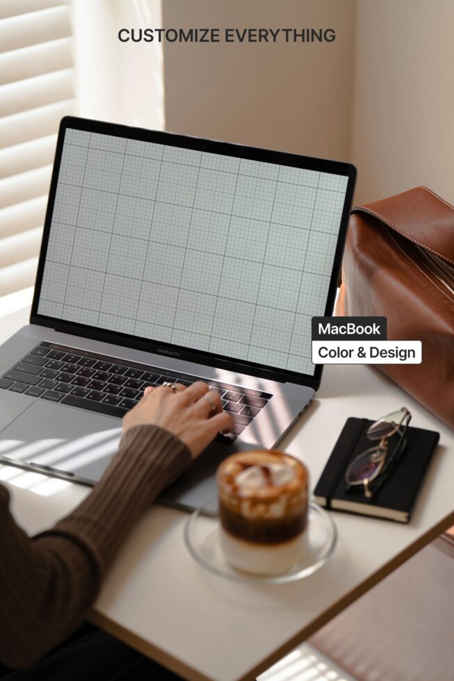 MacBook with blank grid screen on desk next to leather bag, coffee, notebook