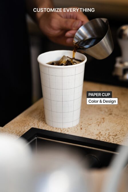 Barista pouring espresso into gridded paper cup with metal milk pitcher mid-pour