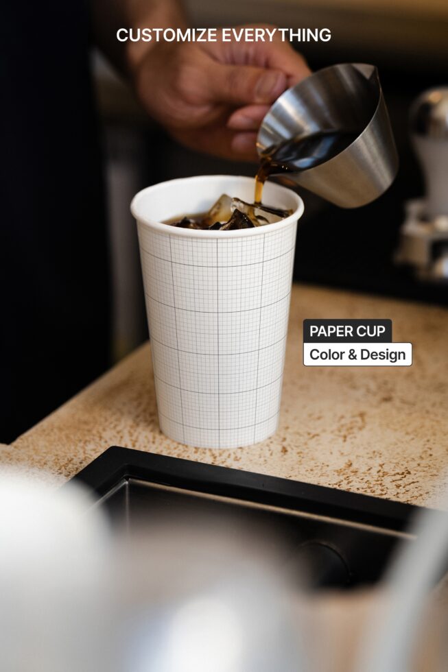 Barista pouring espresso into gridded paper cup with metal milk pitcher mid-pour