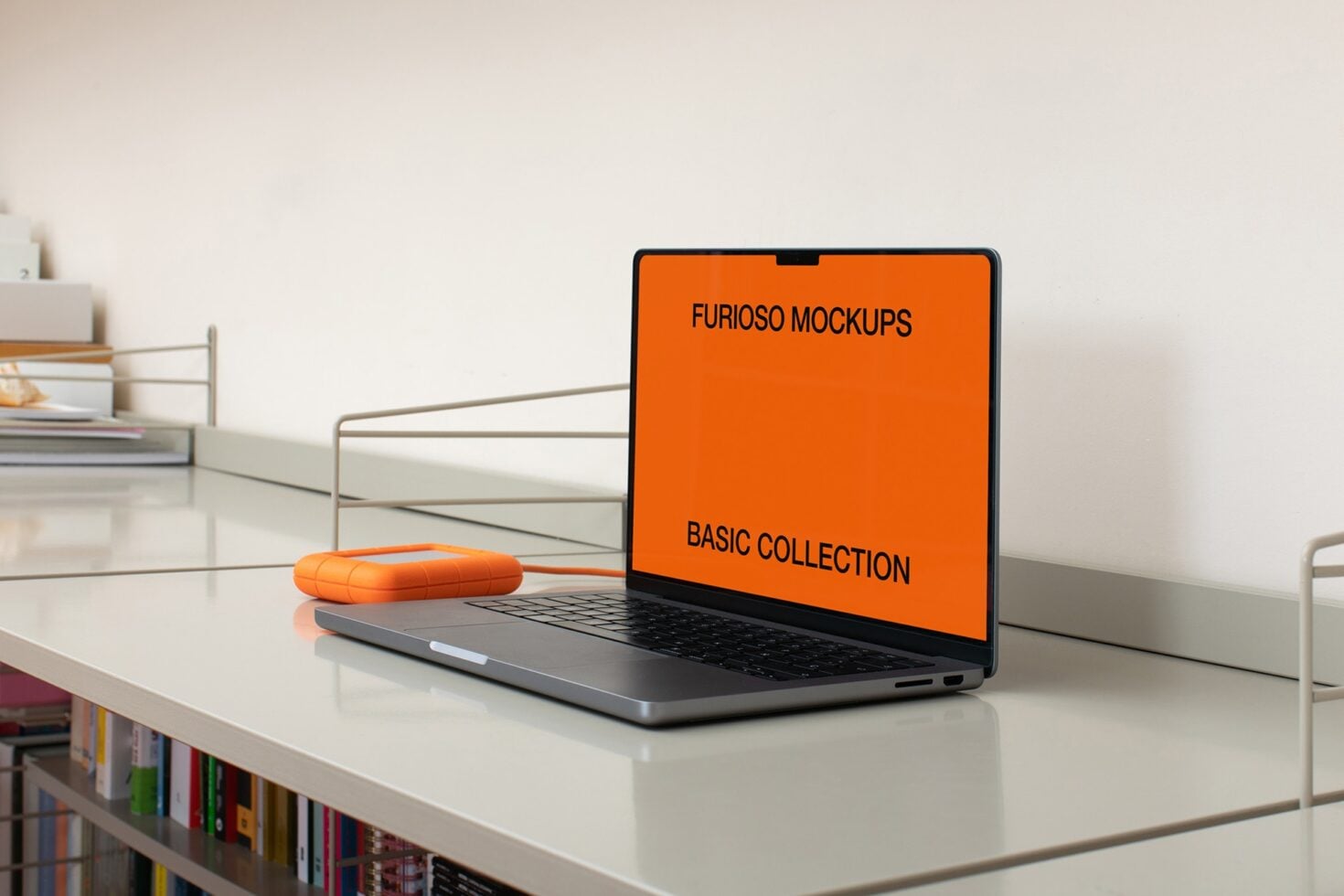 MacBook on white desk with orange mouse next to it, bookshelves in background