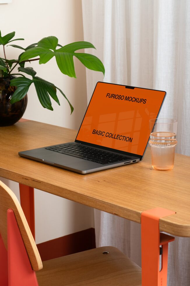 Open laptop on wooden desk with orange screen, plant pot, water glass beside it