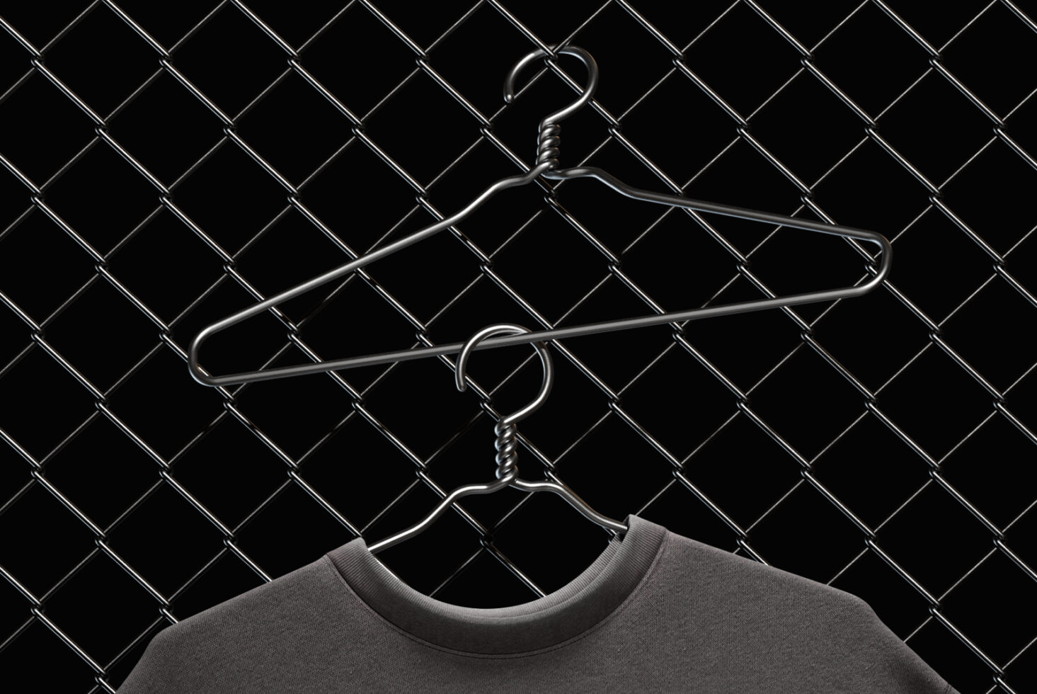 Two metal hangers draped with charcoal tee on chain-link fence backdrop