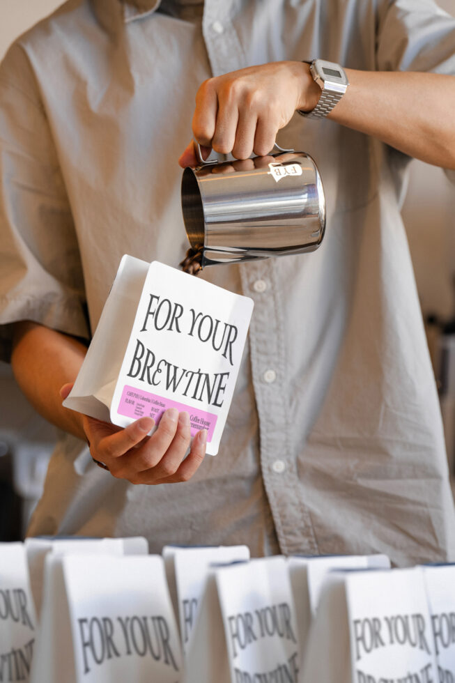 Person pouring from metal pitcher over white coffee bag mockup with pink label
