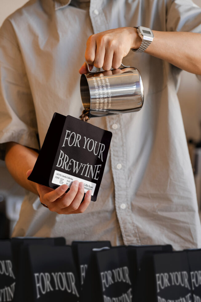 Person holding metal milk pitcher over black coffee bag mockup with white label