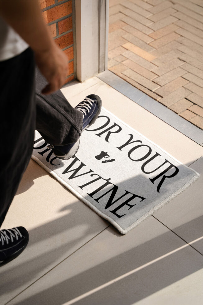 Person stepping on doormat with dry your wine text, black sneakers on entryway