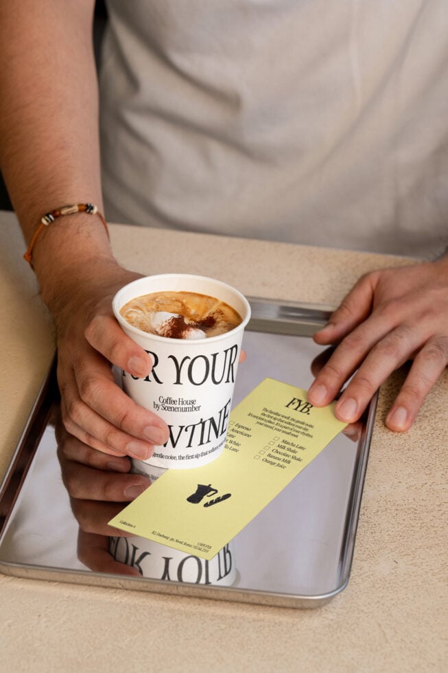Hands holding branded cup with latte over tray, yellow menu card visible below