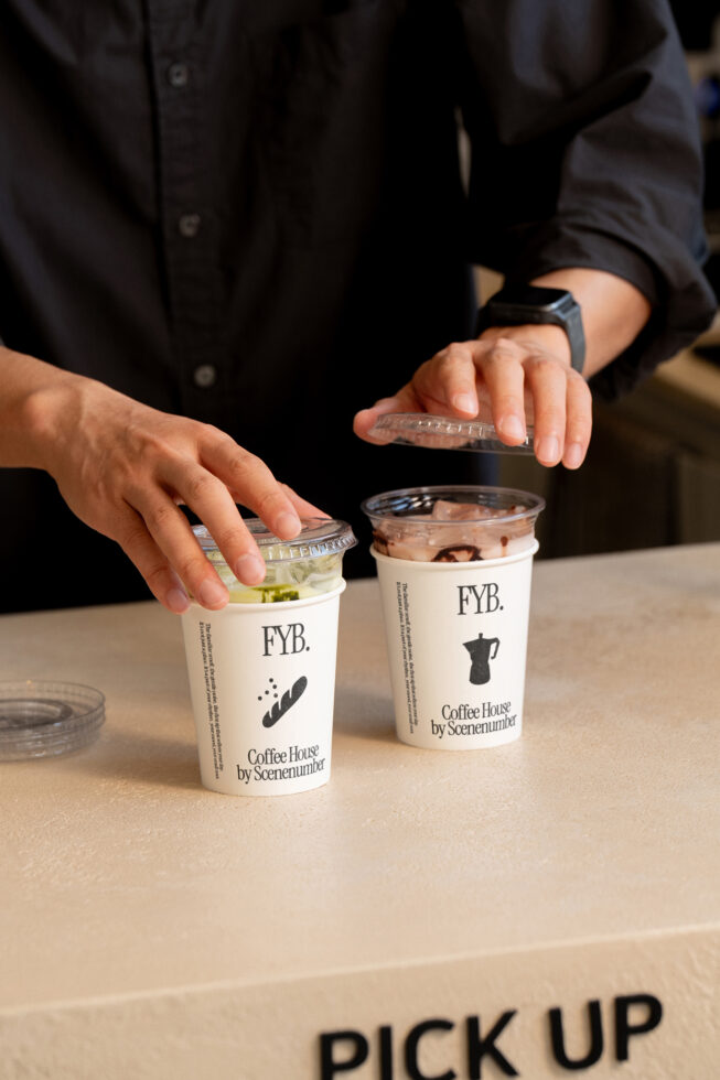 Hands holding two branded coffee cups with clear lids, one with matcha drink