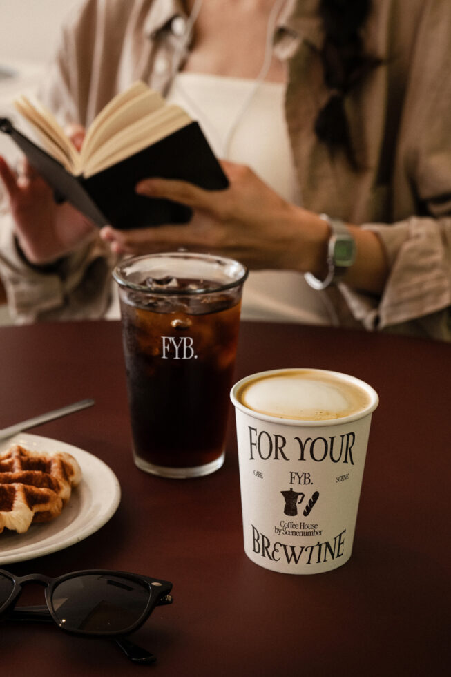 Person reading book with yellow coffee cup, iced drink, waffles on dark table