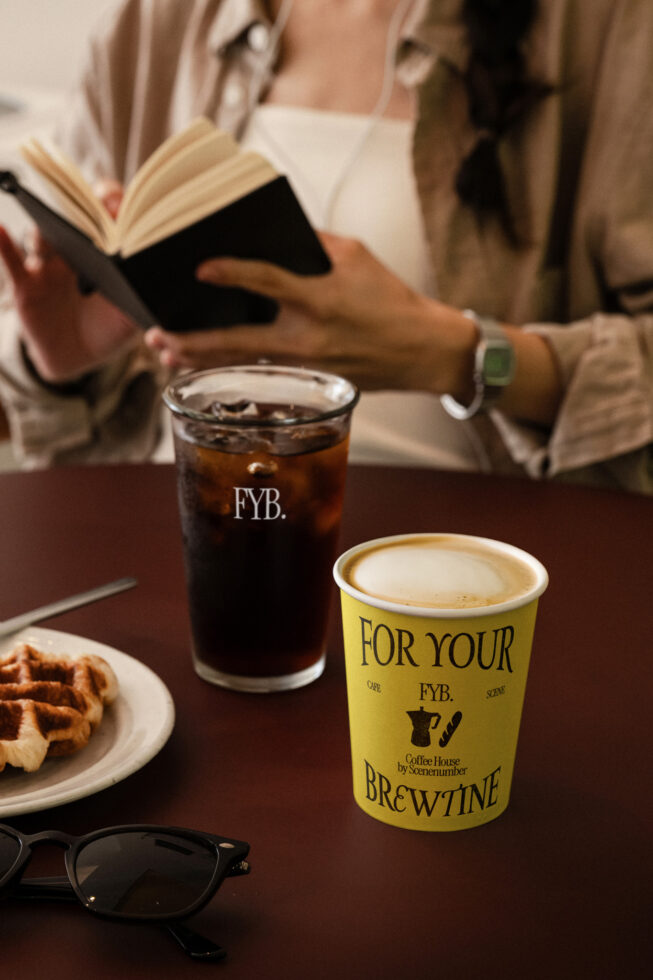 Yellow coffee cup with latte, iced drink, book, waffles on dark wood table
