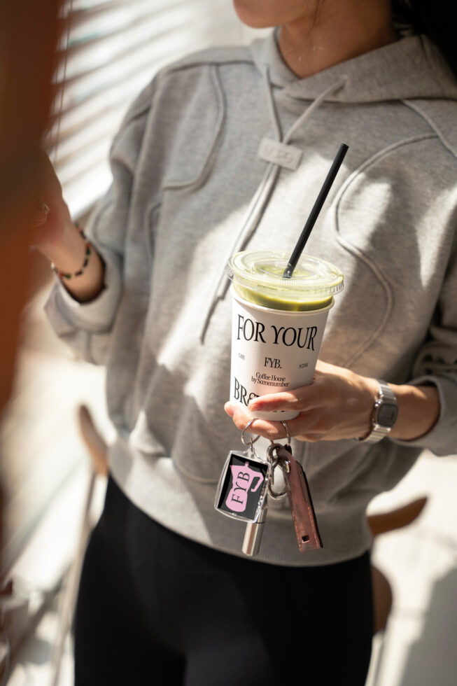Person in beige hoodie holding iced matcha drink with keychain hanging from cup