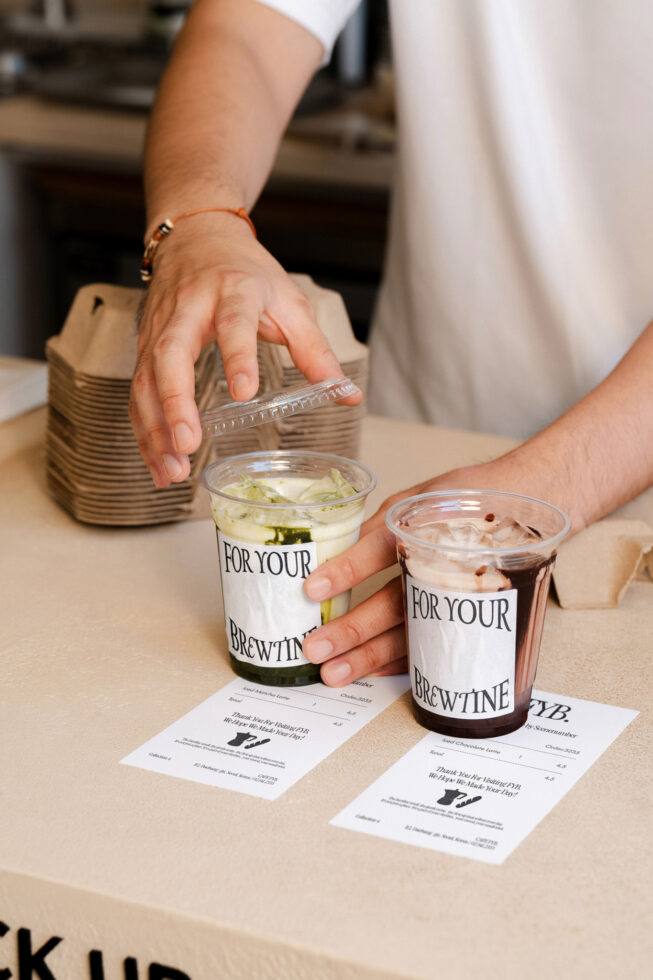 Hands holding two paper cups with For Your Brewtine labels, receipts on counter