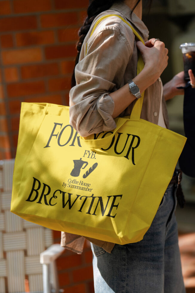 Bright yellow tote bag featuring For Your Brewtine coffee house design carried on person by brick wall